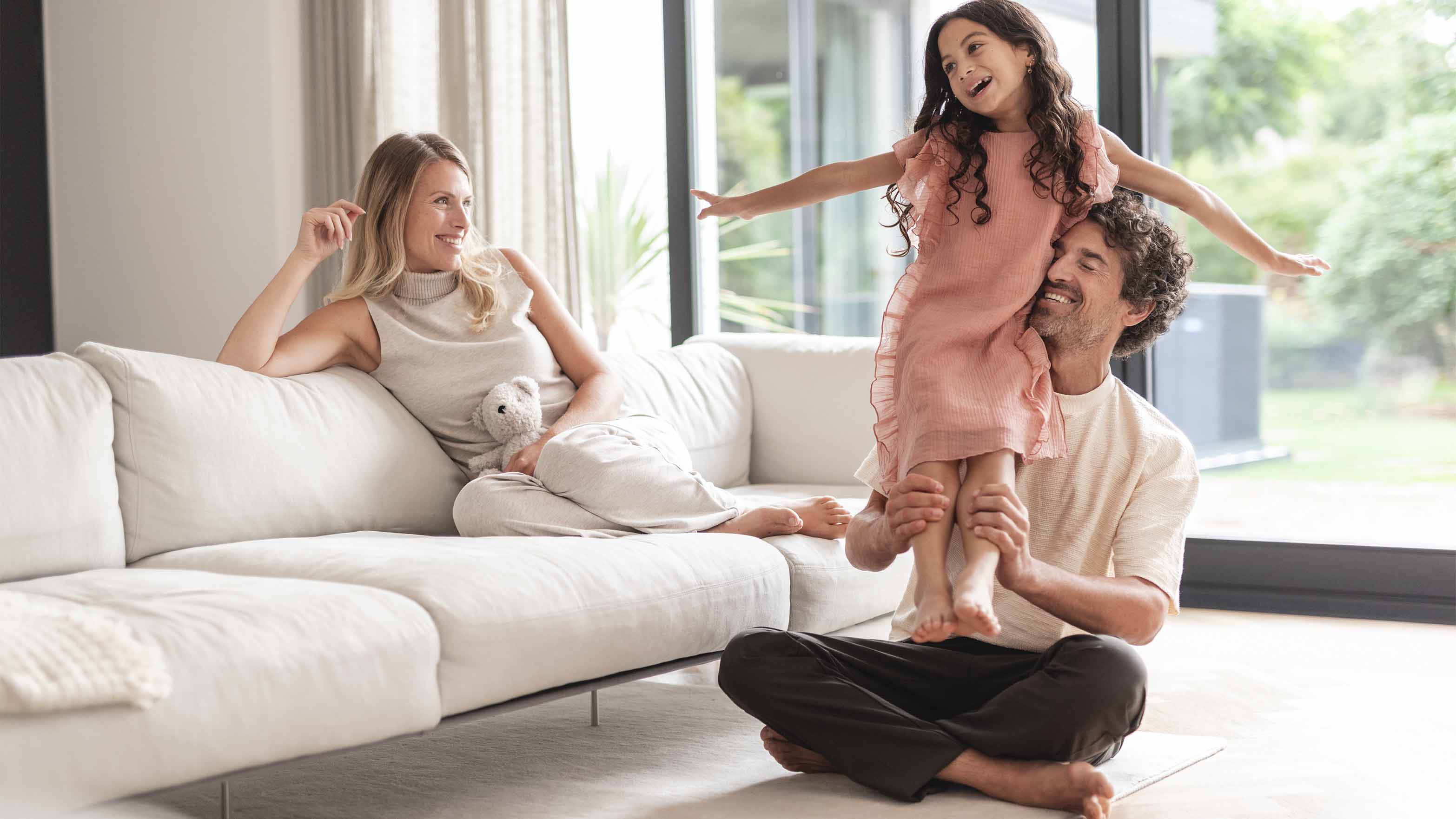 Eine Familie spielt im Wohnzimmer. Im Hintergrund ist eine Wärmepumpe im Garten zu sehen.