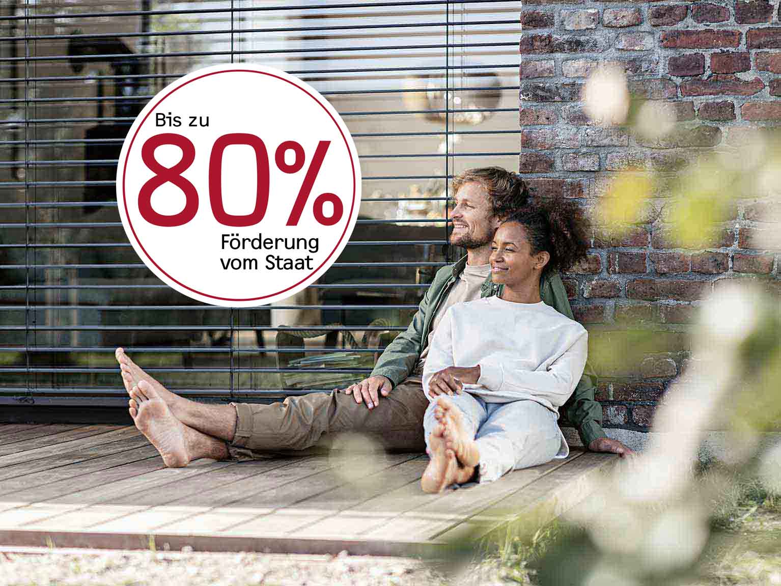 Mann und Frau sitzen barfuß auf dem Boden ihrer Terasse, ein rundes Signet mit weißem Hintergrund zeigt Jetzt bis zu 70% Förderung vom Staat
